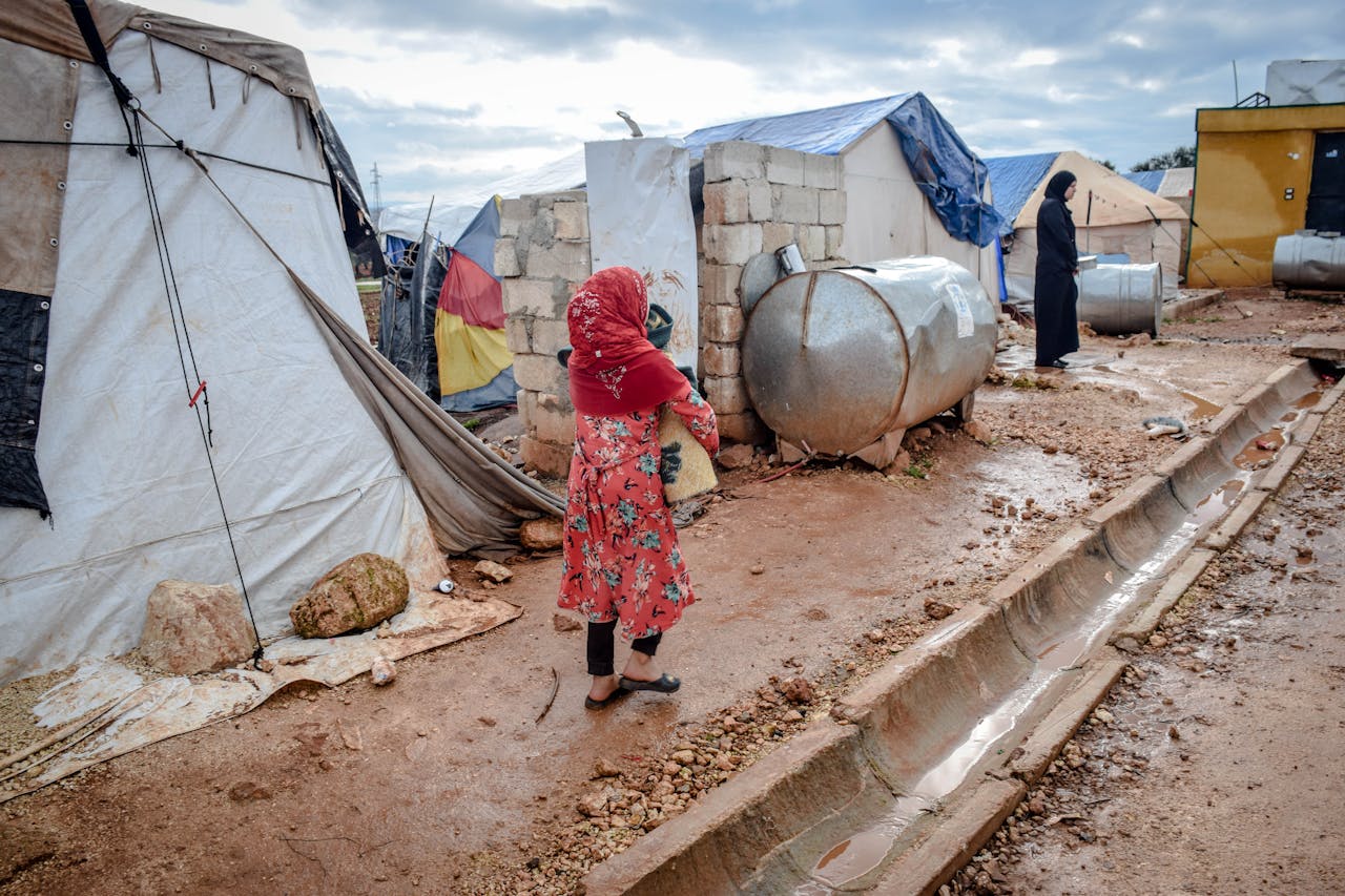 Two women walk through a refugee camp in Idlib, Syria, against a cloudy sky backdrop.